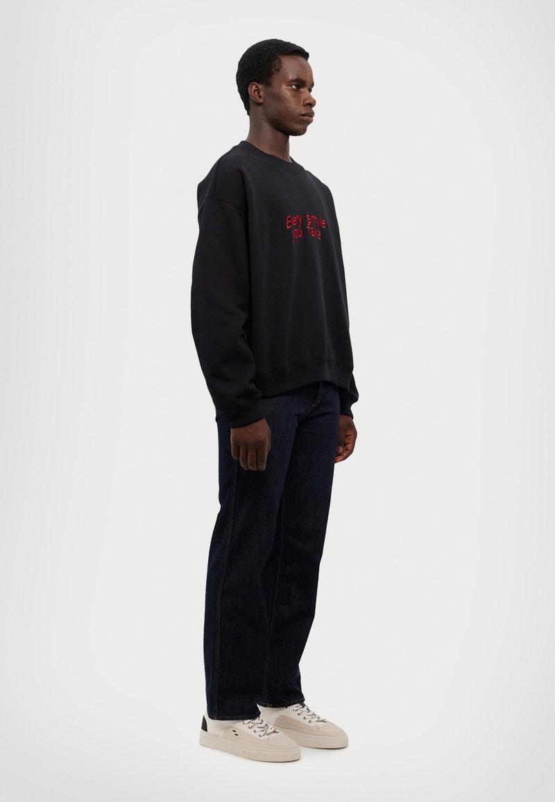 Felpa nera con scritta rossa che recita "Every Smile You Take", abbinata a jeans di denim blu scuro e sneakers bianche con dettagli neri.