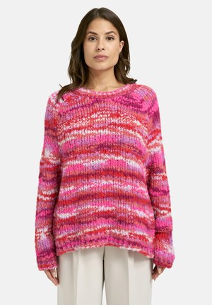 Frau in einem grob gestrickten Pullover mit horizontalen Streifen in Rosa-, Rot- und Weißtönen, kombiniert mit hellen, weiten Hosen.
