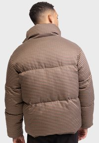 Doudoune marron à carreaux avec un col haut. Présente une texture matelassée et une coupe décontractée. Couverture complète avec une taille cintrée.