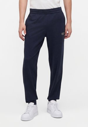Man draagt donkerblauwe joggingbroek met elastische taille en witte sneakers, staand tegen een effen witte achtergrond.