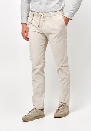 Man draagt beige slim-fit broek met trekkoord in de taille en bijpassende beige sneakers, staand tegen een effen achtergrond.
