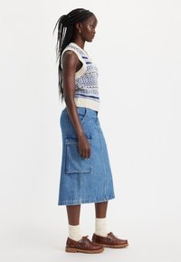 Strikket vest med blå og creme mønstre, høj halsudskæring og korte ærmer, parret med en lang denimnederdel med en lomme og frynset kanten.