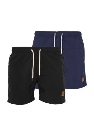 Homme portant un short bleu marine à cordon et des sandales noires à enfiler, debout avec une main dans la poche.