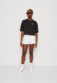 T-shirt cropped nera con logo Puma, pantaloni corti bianchi con scritta "PUMA" e sneakers bianche con plateau, il tutto abbinato a occhiali da sole neri.