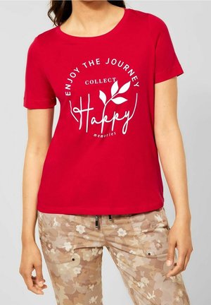 Kvinde iført en rød t-shirt med hvid tekst "Enjoy the journey, collect happy memories" og beige bukser med blomstermønster.
