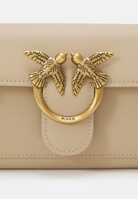 Bolsa de mão em couro bege, com um hardware redondo em tom dourado e dois apliques de pássaros. Textura suave com detalhes de costura visíveis.