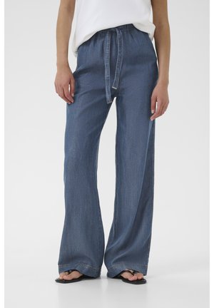 Szerokie niebieskie spodnie dżinsowe z szerokimi nogawkami i ściągaczem w pasie, noszone z czarnymi sandałami, prezentowane na osobie stojącej na jednolitym tle.