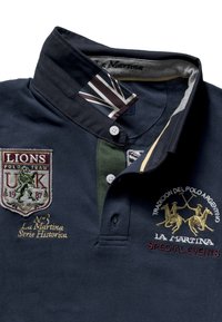 Granatowa koszulka polo z zielonym kołnierzem, z haftowanymi naszywkami, w tym emblematu lwa i grafik drużyny polo. Zapinana na guziki, wykonana z bawełny.