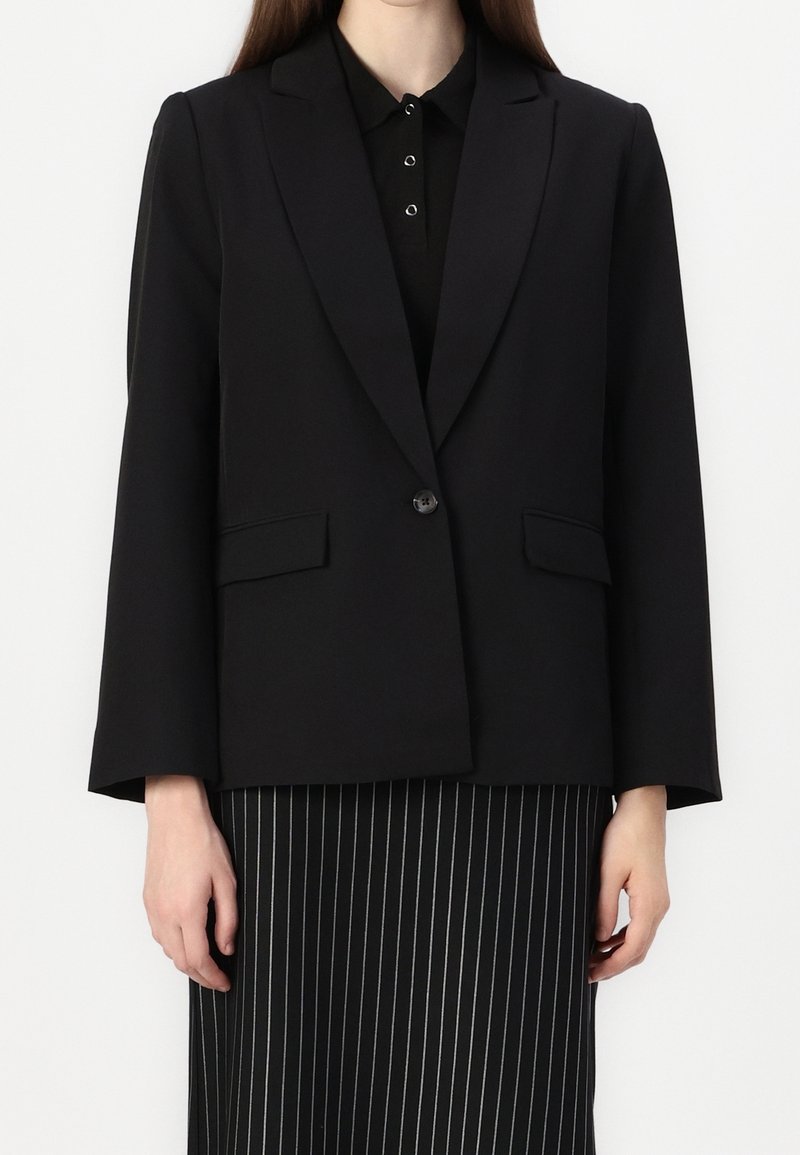 Femme portant un blazer noir à un seul bouton sur une chemise noire à col et une jupe noire à fines rayures verticales blanches.