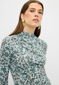 Langärmliges Kleid mit floralem Muster, das blaue und weiße Blumen auf einem braunen Hintergrund zeigt. Hoher Kragen und geraffte Textur. Goldene Statement-Ohrringe.