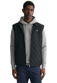 Black quilted vest with sleeveless design, worn over a gray hoodie. Features a zip front and small logo on the left chest.