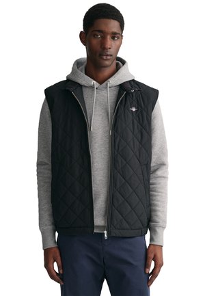 Black quilted vest with sleeveless design, worn over a gray hoodie. Features a zip front and small logo on the left chest.