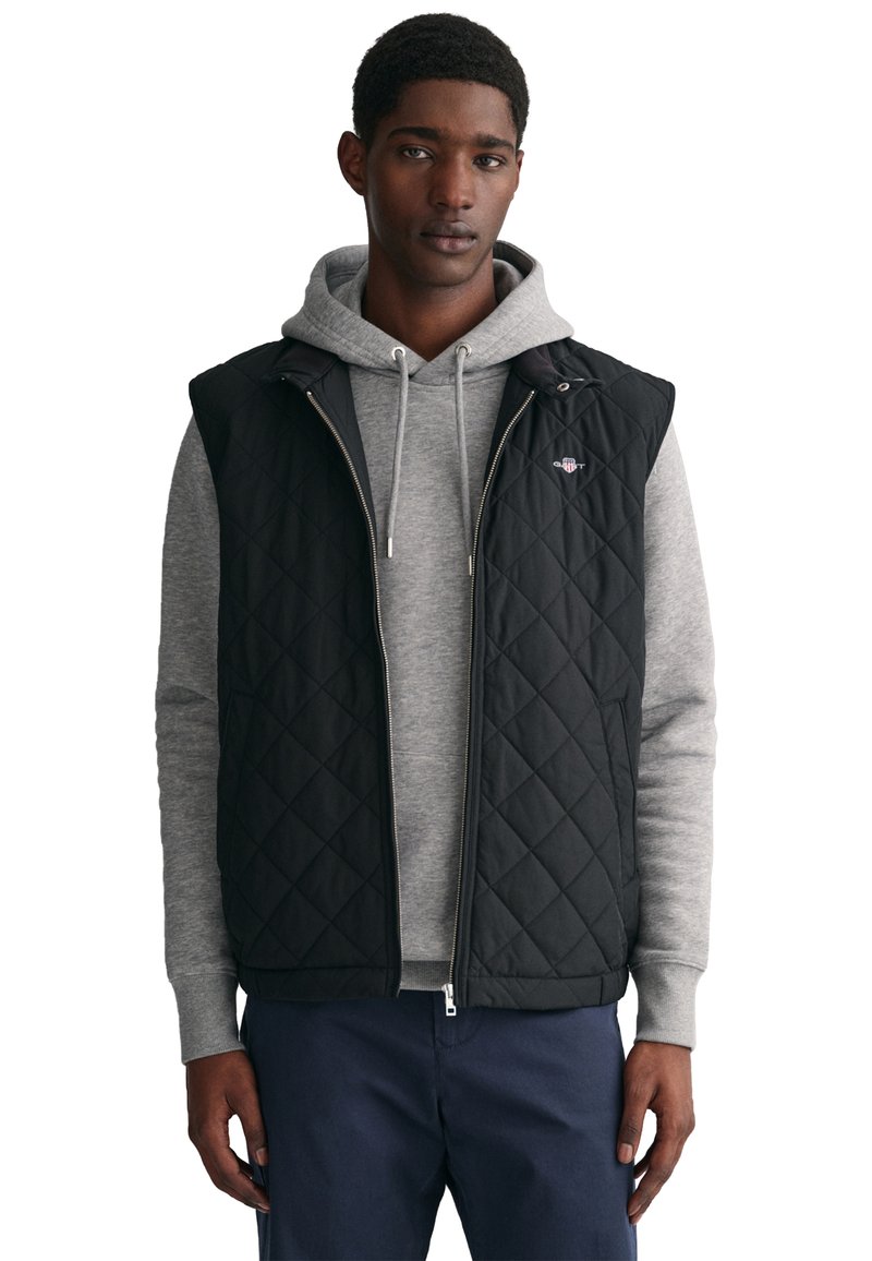 Gilets noir matelassé à design sans manches, porté par-dessus un sweat à capuche gris. Comprend une fermeture éclair à l'avant et un petit logo sur la poitrine gauche.