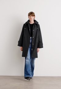 Black hooded coat with a relaxed fit, featuring snap closures, front pockets, and adjustable drawstrings. Worn over a black top and blue jeans.