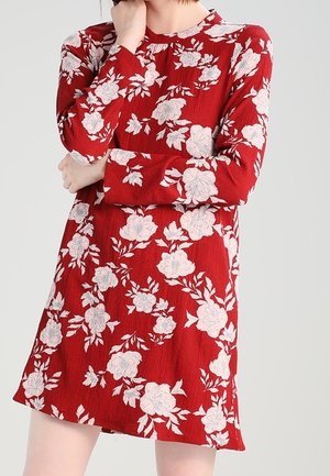 Femme portant une robe rouge mi-longue à manches longues avec un motif floral blanc, debout les bras croisés et une main touchant son visage.
