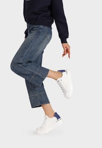 Sneaker bianchi con dettagli blu, tomaia in pelle liscia, design con lacci e suola in gomma spessa, abbinati a pantaloni in denim a vita bassa.