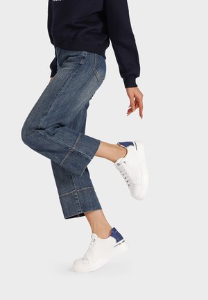 Sneaker bianchi con dettagli blu, tomaia in pelle liscia, design con lacci e suola in gomma spessa, abbinati a pantaloni in denim a vita bassa.