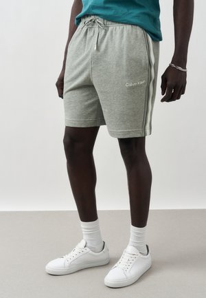 Hombre que lleva pantalones cortos grises Calvin Klein con franjas laterales, calcetines blancos, zapatillas blancas y una camiseta verde azulado, de pie en el interior sobre un suelo gris.