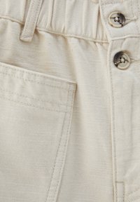 Gros plan sur un pantalon beige montrant une ceinture élastique, une poche avant et deux boutons marron sur la braguette.