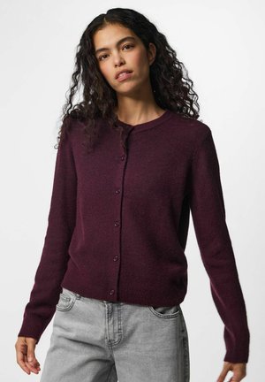 Femme aux longs cheveux bouclés portant un cardigan violet foncé boutonné et un jean gris clair, debout devant un fond uni.