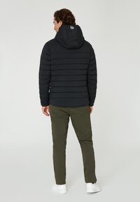 Svart puffjacka med horisontella quiltade sektioner, anpassad huva och liten cirkulär logotyp på baksidan. Bärs med olivgröna byxor och vita sneakers.