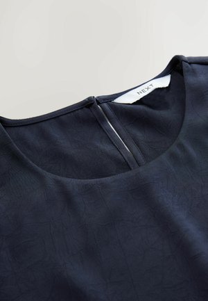 Blusa azul marino con un escote redondo, tejido texturizado y cremallera en la parte posterior. La etiqueta dice "NEXT". Acabado suave con un caído blando.