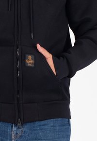 Mano in tasca di una felpa nera con zip e patch del logo, abbinata a jeans blu su uno sfondo bianco.