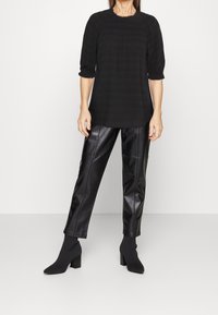 Blouse noire texturée avec encolure froncée, associée à un pantalon en simili cuir noir et des bottines à chaussettes. Toutes les pièces ont une coupe ajustée.