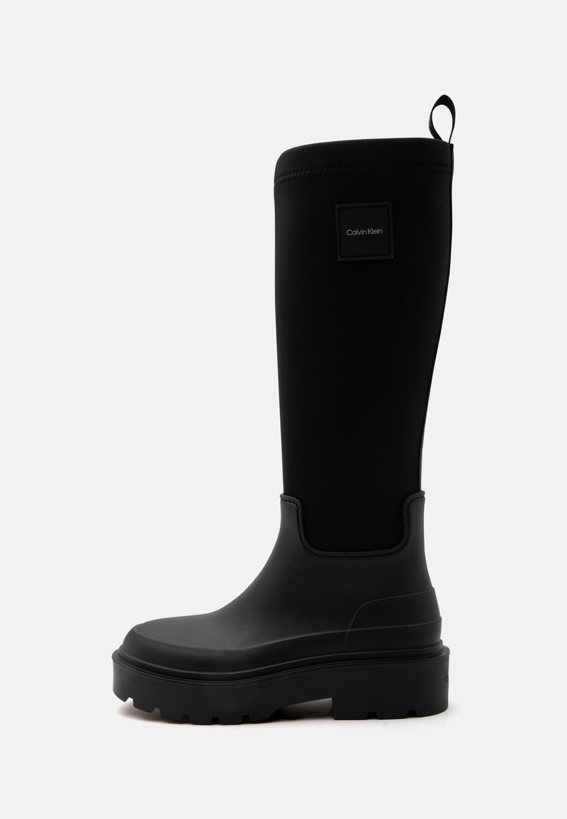 Botte haute en caoutchouc noir avec une semelle à crampons épaisse, languette à l'arrière et petit logo carré Calvin Klein près du haut.