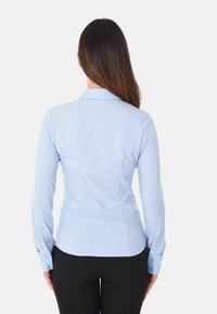 Camicia a maniche lunghe di colore blu chiaro con design aderente, con polsini bottonati e colletto. Tessuto liscio con sottile texture, indossata con pantaloni neri.