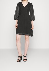 Svart klänning med V-ringning, knapprad och genomskinliga långa ärmar. Texturerad tyg med prickigt mönster och volangdetaljer. Svarta klacksandaler.