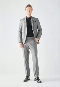 Giovane uomo in piedi, con un completo grigio chiaro, una camicia nera e scarpe eleganti nere, davanti a uno sfondo chiaro e semplice.