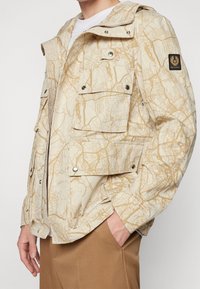 Beige jacka med kartmönster, som har flera fickor, tryckknappar och huva. Tyget verkar vara lätt och strukturerat.