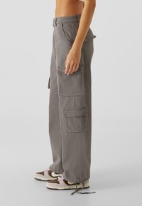 Pantalons cargo gris en coton durable avec une coupe ample, dotés de poches latérales et de poignets ajustables. Portés avec des baskets marron et rose.