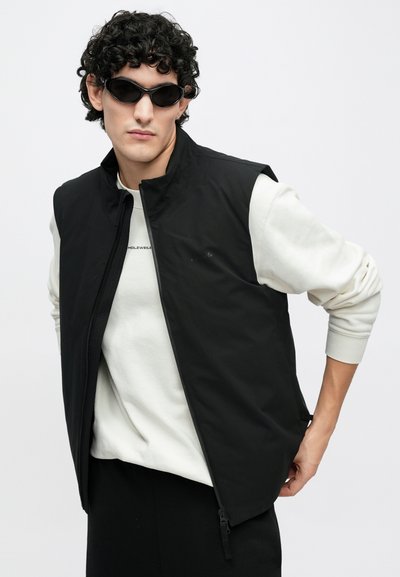 Fekete, ujjatlan mellény magas nyakkal, cipzáras záródással és oldalsó cipzárakkal, fehér logós pulóver fölött viselve. Fekete napszemüveg egészíti ki a megjelenést.