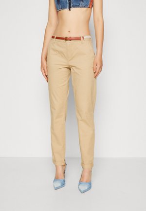 Kvinde iført beige opkædte bukser med et rødt og hvidt bælte, blå spidse hæle og en kort denimtop.