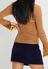 Prążkowany, złoto-brązowy top z długim rękawem i rozkloszowanymi rękawami zdobionymi żółtymi falbankami, w połączeniu z granatową, sztruksową mini spódniczką.