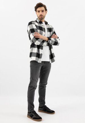 Junger Mann steht mit verschränkten Armen, trägt ein schwarz-weiß kariertes Hemd, ein weißes T-Shirt, graue Jeans und schwarze Turnschuhe.