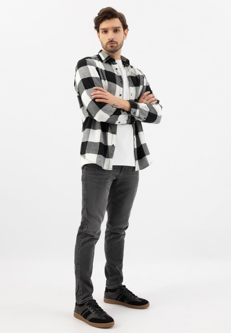 Giovane in piedi con le braccia incrociate che indossa una camicia a quadretti bianca e nera, una t-shirt bianca, jeans grigi e sneaker nere.