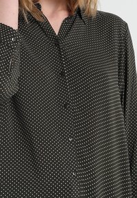 Camicia button-up verde scuro con pois bianchi, maniche lunghe e collo. Il tessuto appare liscio con una leggera lucentezza.