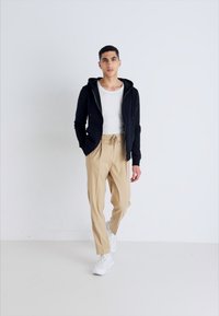 Juodas zip-up džemperis, baltas marškinėlis, smėlio spalvos kelnės ir baltos sportbačiai. Modelis stovi prieš neutralų foną, demonstruodamas aprangą.