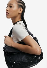 Schwarze Ledertasche mit silbernen Nieten und Nähdetails, getragen über einem beigen T-Shirt; verfügt über einen Klappenverschluss und einen verstellbaren Trageriemen.
