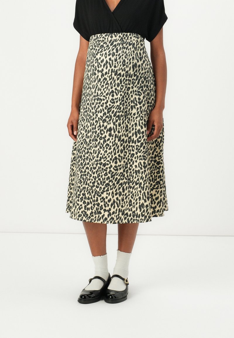 Femme portant un haut noir et une jupe midi beige avec un imprimé léopard noir, associée à des chaussettes blanches et des chaussures Mary Jane noires.