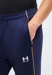 Άτομο που φοράει αθλητικό παντελόνι Under Armour σε ναυτικό μπλε χρώμα με ελαστική μέση, πορτοκαλί πλευρική ρίγα και χέρι σε τσέπη με φερμουάρ.