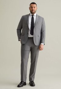 Grijze tweedelige kostuum bestaande uit een op maat gemaakt jasje en vlakke broek, gecombineerd met een wit shirt en een zwarte stropdas. Zwarte schoenen maken de look af.