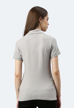 Frau mit langen braunen Haaren, die ein eng anliegendes hellgraues kurzärmeliges Polo-Shirt trägt, von hinten vor einem einfarbigen Hintergrund gesehen.