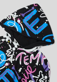 Felpa nera con testo e schizzi di vernice in stile graffiti nei colori blu, viola, rosa e bianco, caratterizzata da forme astratte e lettere stilizzate.