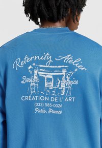 Niebieski sweatshirt z białym graficznym napisem, w tym "Reternity Atelier" oraz szczegółami Paryża, z artystycznym rysunkiem sceny kawowej.