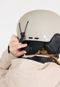 Beige Skihorn mit matter Oberfläche, verstellbarem Kinnriemen und schwarzen Ohrpolstern. Verfügt über Belüftungsöffnungen und Logos an der Seite.