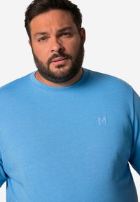 Světle modrý crewneck svetr vyrobený z měkké látky, s jemně vyraženým logem na levé straně hrudi. Hladká textura, regular fit.